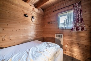 Puuinen mökkihuone, jossa on sänky, ikkuna, jossa on verho, ja seinälle asennettu lämmitin.