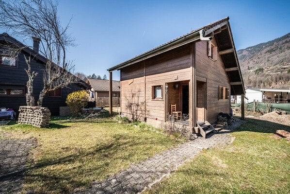 A wooden cabin with a sloped roof, a small porch, and a brick pathway leading to the entrance. - Chalet “6p Classique - Le Rhodo” with Mountain View, Private Terrace, and Wi-Fi (Verchaix)