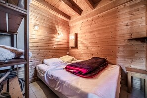 A wooden cabin room with a bed, a shelf with a fluffy item, and a wall-mounted light fixture. - Chalet “6p Classique - Le Rhodo” with Mountain View, Private Terrace, and Wi-Fi (Verchaix)