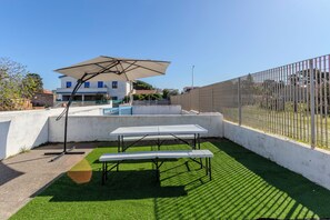 Eine Terrasse mit Tisch und Sonnenschirm, umgeben von einem weißen Zaun und einem Gebäude im Hintergrund.