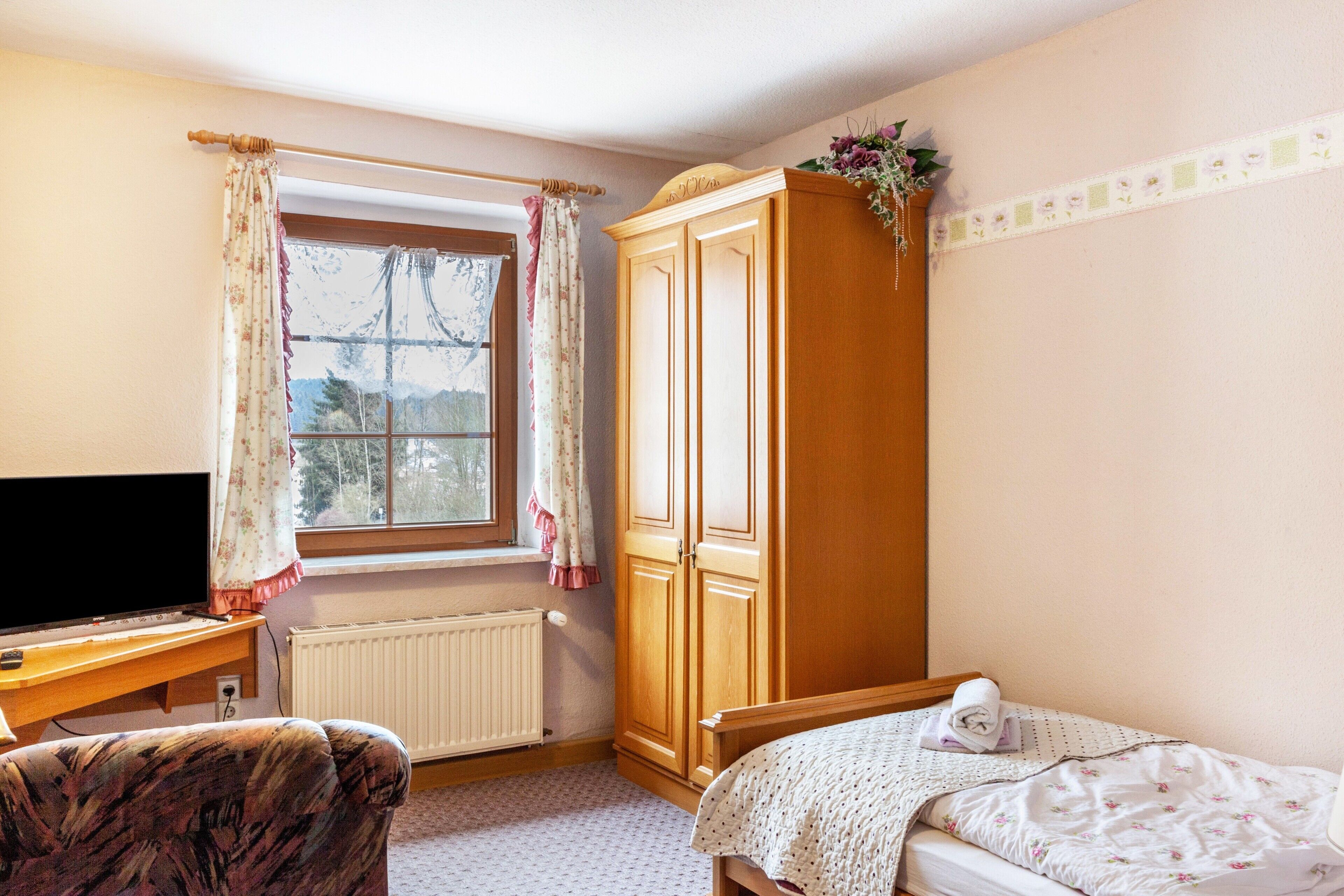 A bedroom with a bed, a wooden wardrobe, a desk with a television, and a window with curtains.
