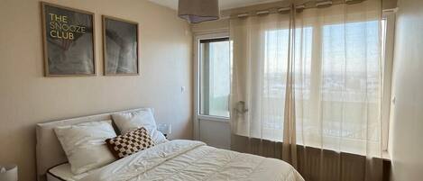 A bedroom with a bed, a nightstand, a lamp, a window with sheer curtains, and two wall art pieces.