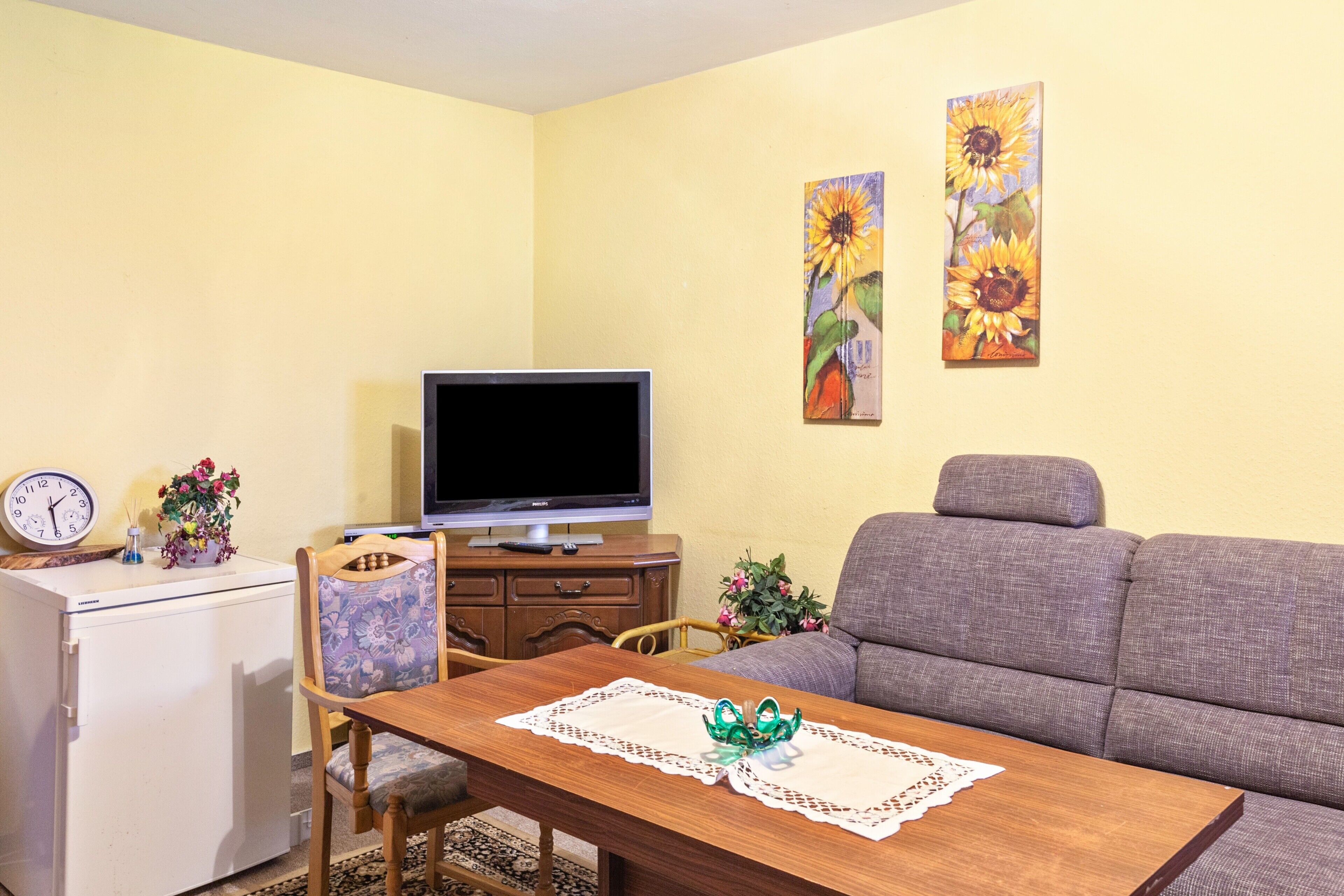 Una sala de estar con televisión, sofá, mesa de comedor y un reloj sobre una mesita.