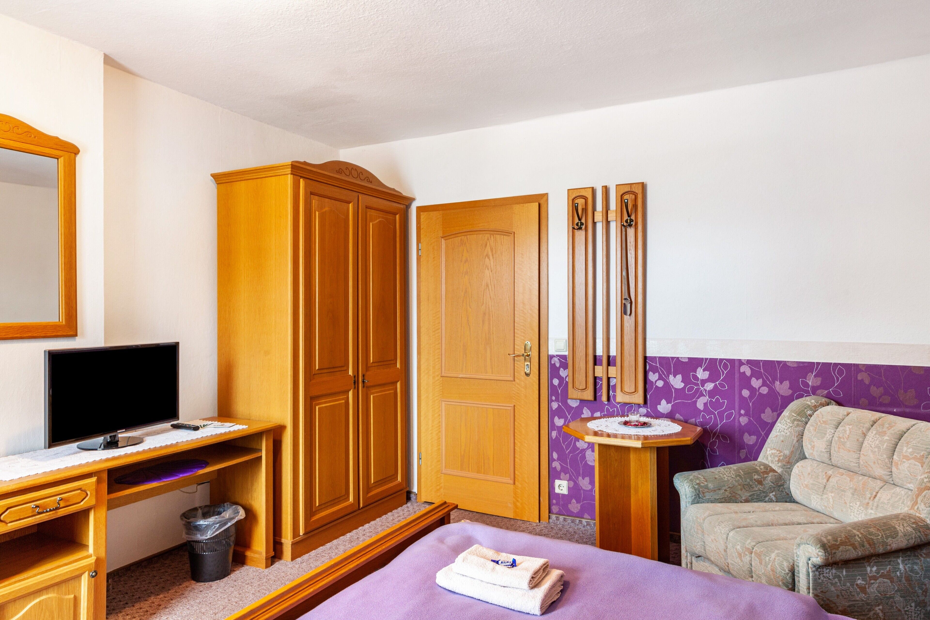 Una habitación con cama, escritorio con computadora, armario de madera, espejo y pared morada.