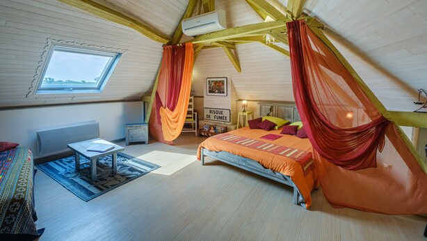 A high-ceilinged room with a sloped ceiling, a bed with orange bedding, a small table, a chair, a window, and a radiator. - Le Balcon De Loire - Curry Room: Escape and Serenity by the Loire (La Chapelle sur Loire)