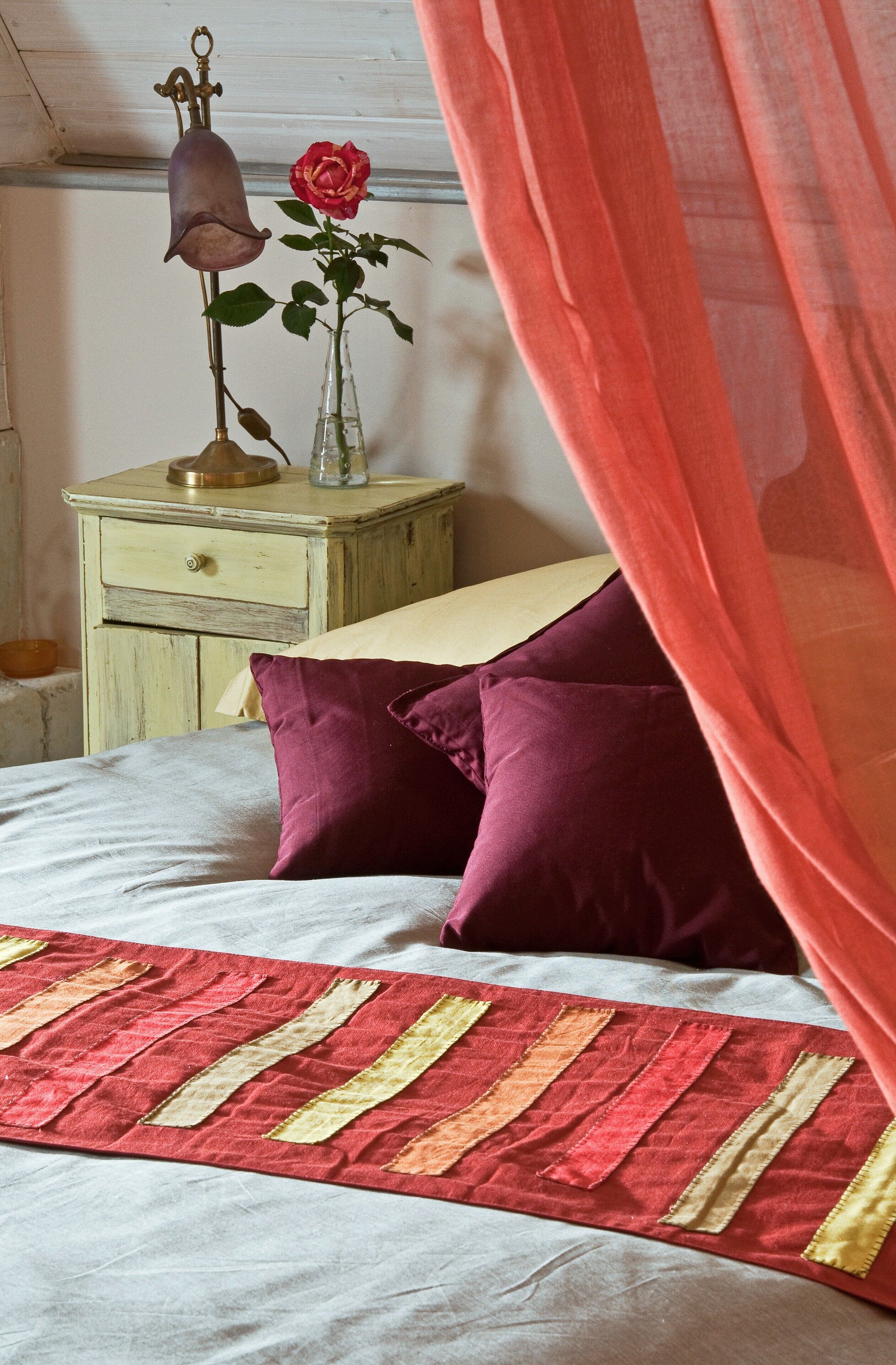 A bed with a red and beige striped bedspread, two purple pillows, a nightstand with a brass bell and a red rose in a glass vase, and a sheer red curtain partially drawn.