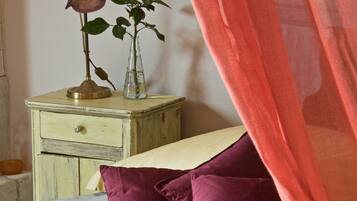 A bed with a red and beige striped bedspread, two purple pillows, a nightstand with a brass bell and a red rose in a glass vase, and a sheer red curtain partially drawn.