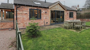 A brick house with a large sliding glass door, a small garden, and a patio with a table and chairs. - Otter's Rest (Droitwich)