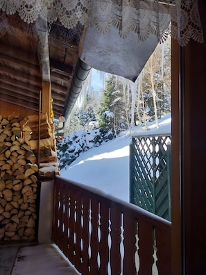 A wooden balcony with a snow-covered landscape view, a stack of firewood, and a decorative lace ceiling.
