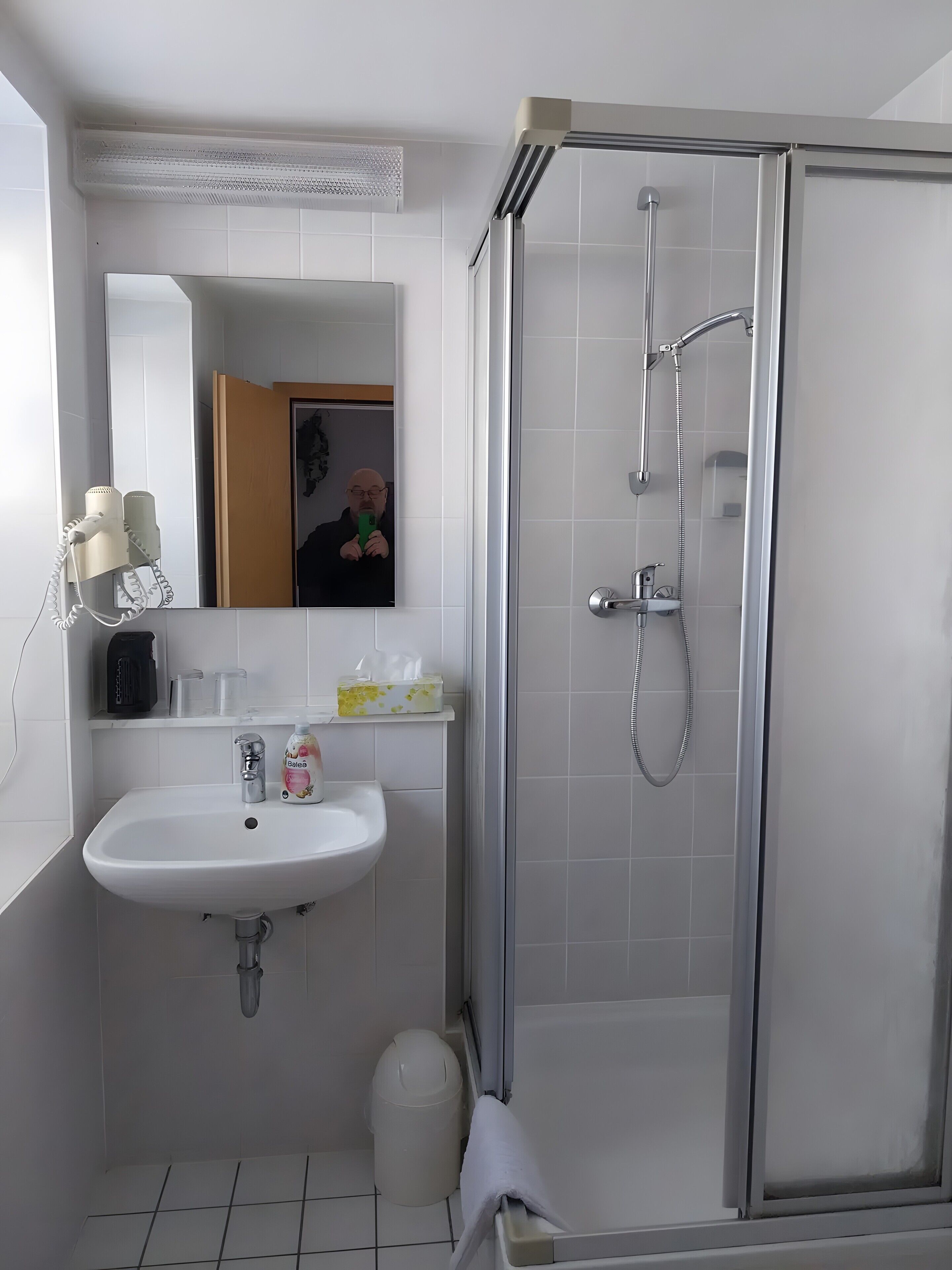 A modern bathroom with a glass-enclosed shower, a white sink with a mirror above it, and a trash bin.