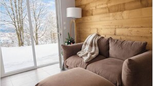 Ett mysigt rum med en brun soffa, en vit lampa och utsikt över snötäckta träd genom ett stort fönster.