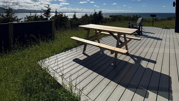 A wooden deck with picnic tables and chairs, overlooking a body of water and distant land. - Lapointe Chalets - Chalet Deveau (Inverness County)