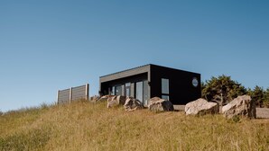 Een modern gebouw met een plat dak, grote ramen en een donkere gevel. Het ligt op een grasheuvel met grote rotsen ervoor.