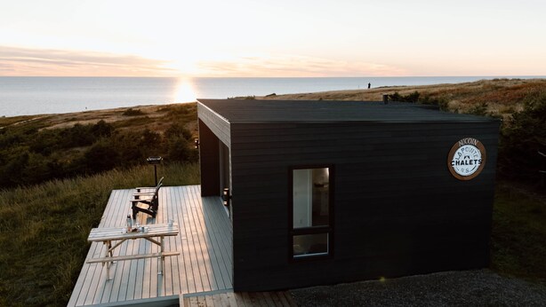 Een klein, donker houten huisje met een terras en uitzicht op zee bij zonsondergang.