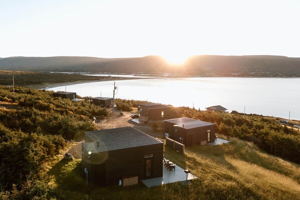 A modern building complex with a flat roof, surrounded by dense vegetation and a body of water in the background.