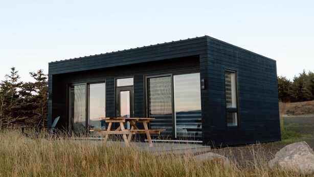 Una cabaña moderna de color oscuro con ventanas grandes y una mesa de picnic al frente.