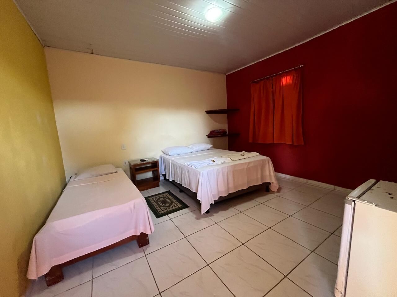A small, clean room with two beds, a small table, a red curtain, and a white refrigerator.