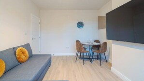Una sala de estar moderna con un sofá, una mesa de comedor con sillas, un reloj en la pared y un televisor de pantalla plana montado.