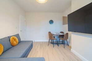 A modern living room with a sofa, a dining table with chairs, a clock on the wall, and a flat-screen TV mounted on the wall. - Modern 2BR Home • Private Driveway • Near Airport (Wythenshawe)