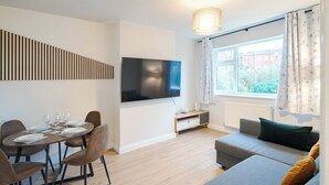 Una sala de estar moderna con un televisor de pantalla plana, una mesa de comedor para cuatro personas, un sofá y una ventana con cortinas.