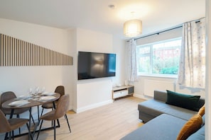 A modern living room with a flat-screen TV, a dining table set for four, a sofa, and a window with curtains. - Modern 2BR Home • Private Driveway • Near Airport (Wythenshawe)