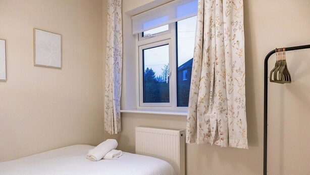 Un dormitorio con cama, ventana con cortinas, radiador y lámpara fijada a la pared.