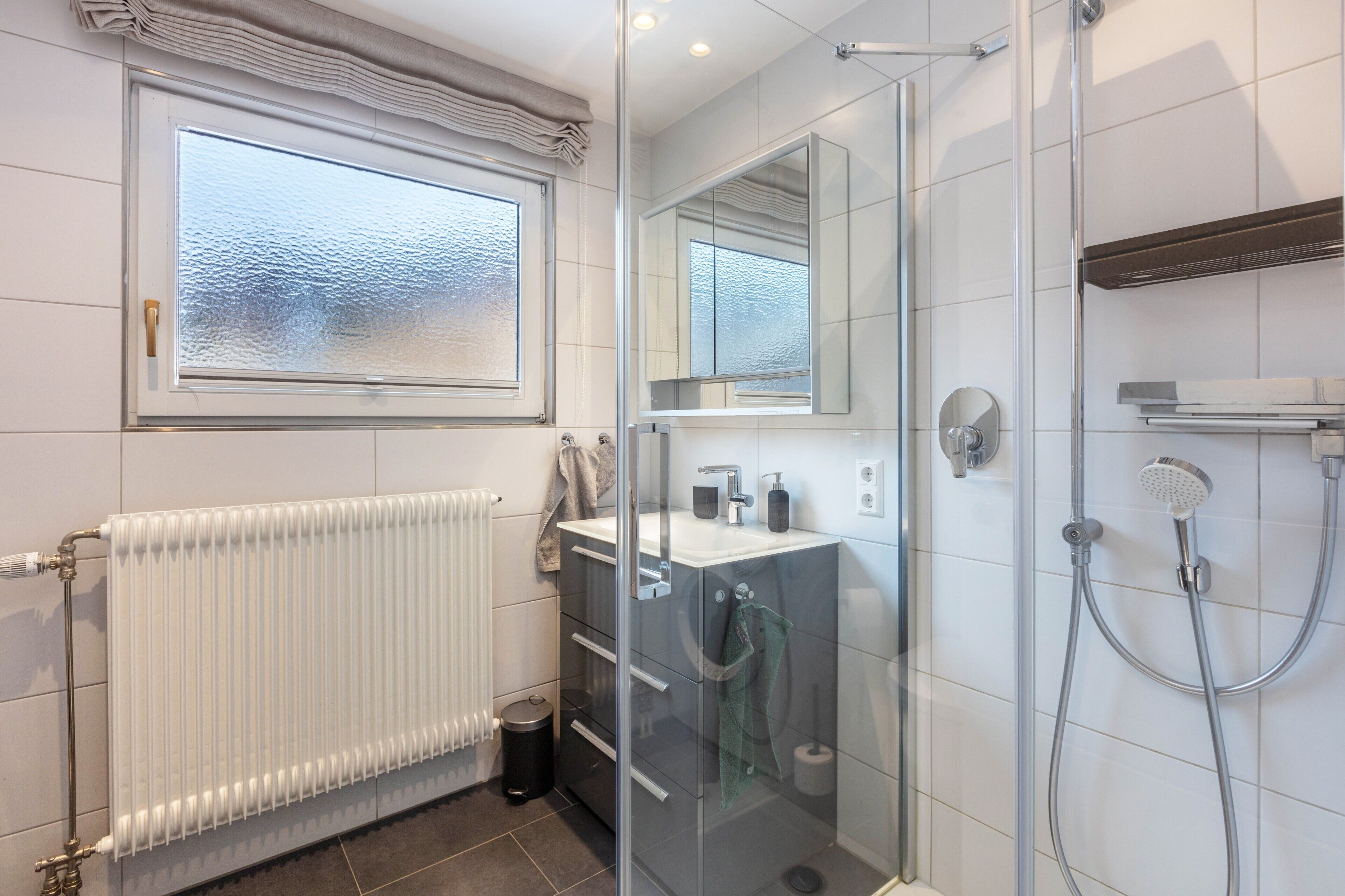 Ein modernes Badezimmer mit Duschkabine aus Glas, einem weißen Handtuchwärmer, einem Waschbecken mit Spiegel und einem Fenster mit Milchglas.