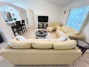 Una sala de estar con un sofá color beige, una mesa de centro, un televisor y una zona de comedor con sillas.