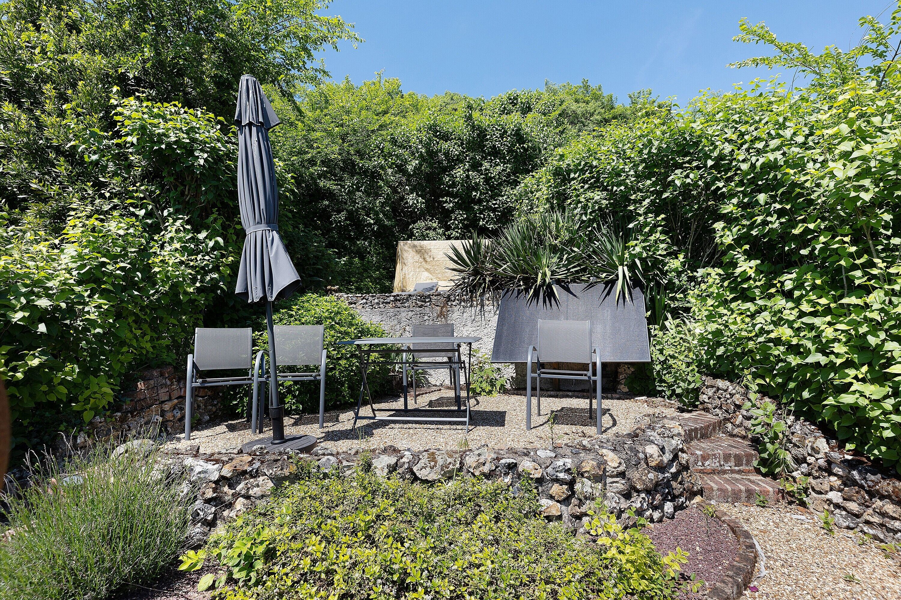 Une terrasse avec une table et des chaises, entourée de verdure et d’un mur en pierre.
