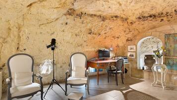 Een kamer met een stenen muur, een bureau met een computer, twee stoelen, een statief en een kleine tafel met een vaas.