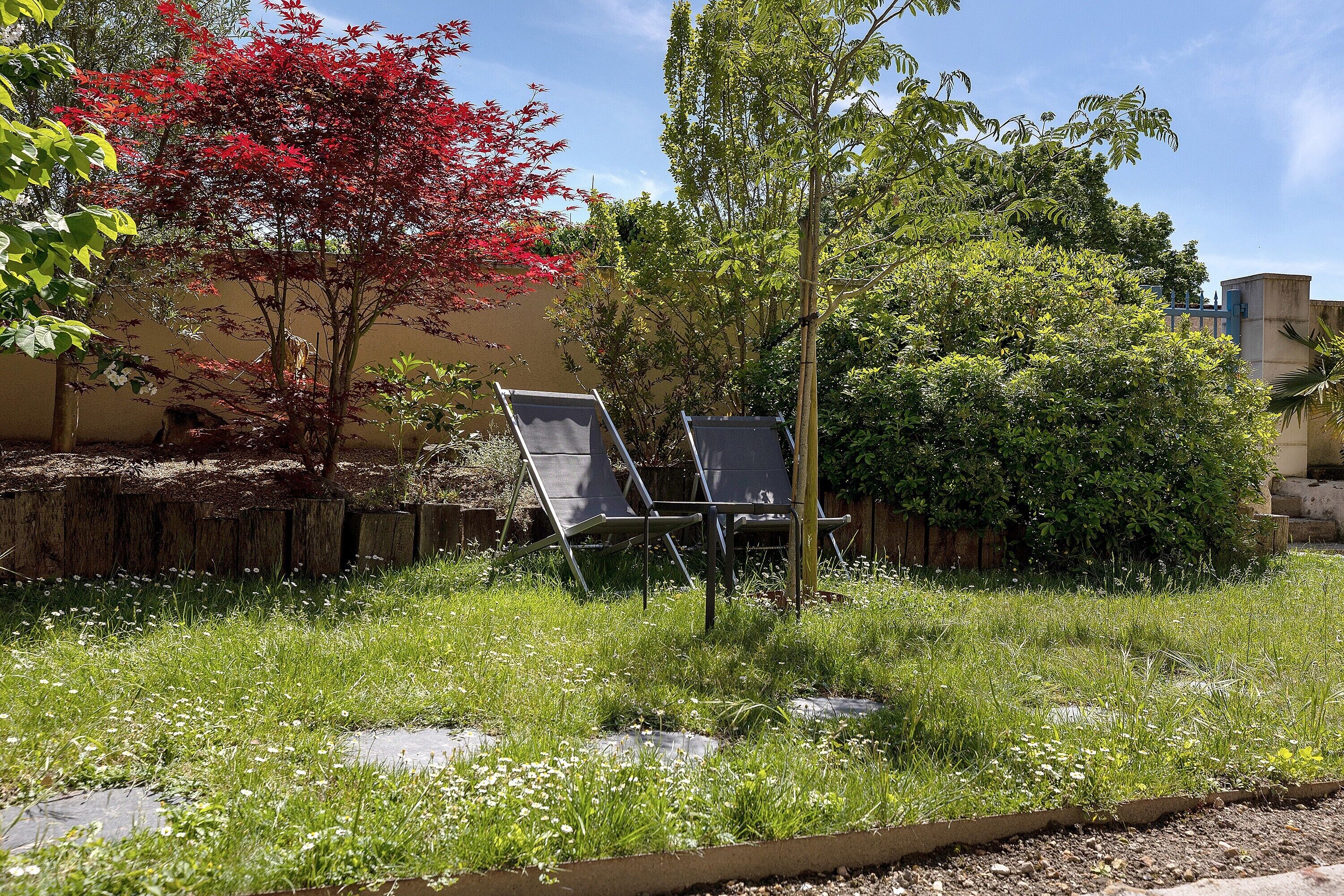 Un jardín con césped, un árbol de hojas rojas y una estructura metálica con sillas.