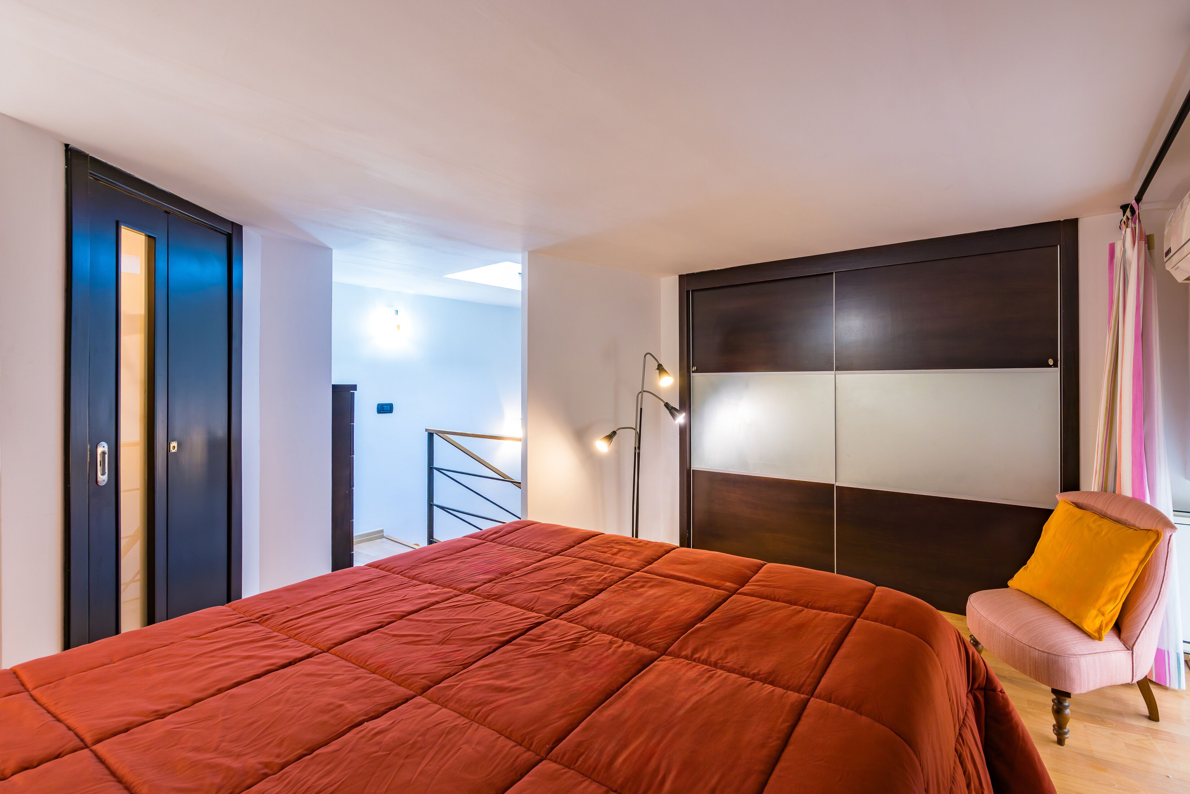 Un dormitorio con una cama grande, un armario, una silla con un cojín amarillo y una escalera al fondo.