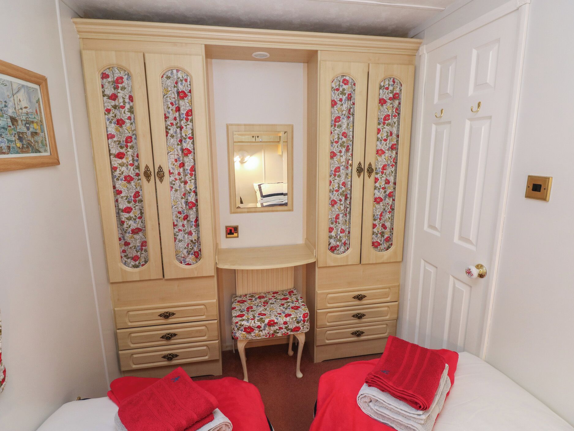 A bedroom with a bed, a small stool, a wooden wardrobe with floral panels, and a mirror above a dresser.
