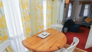 A wooden table with a notebook, surrounded by white chairs in a room with yellow floral curtains and a red carpet. - Rose Villa Caravan (Llandysul)