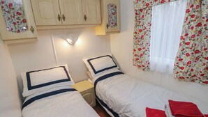 A bedroom with two beds, a wooden wardrobe, and a window with floral curtains. - Rose Villa Caravan (Llandysul)