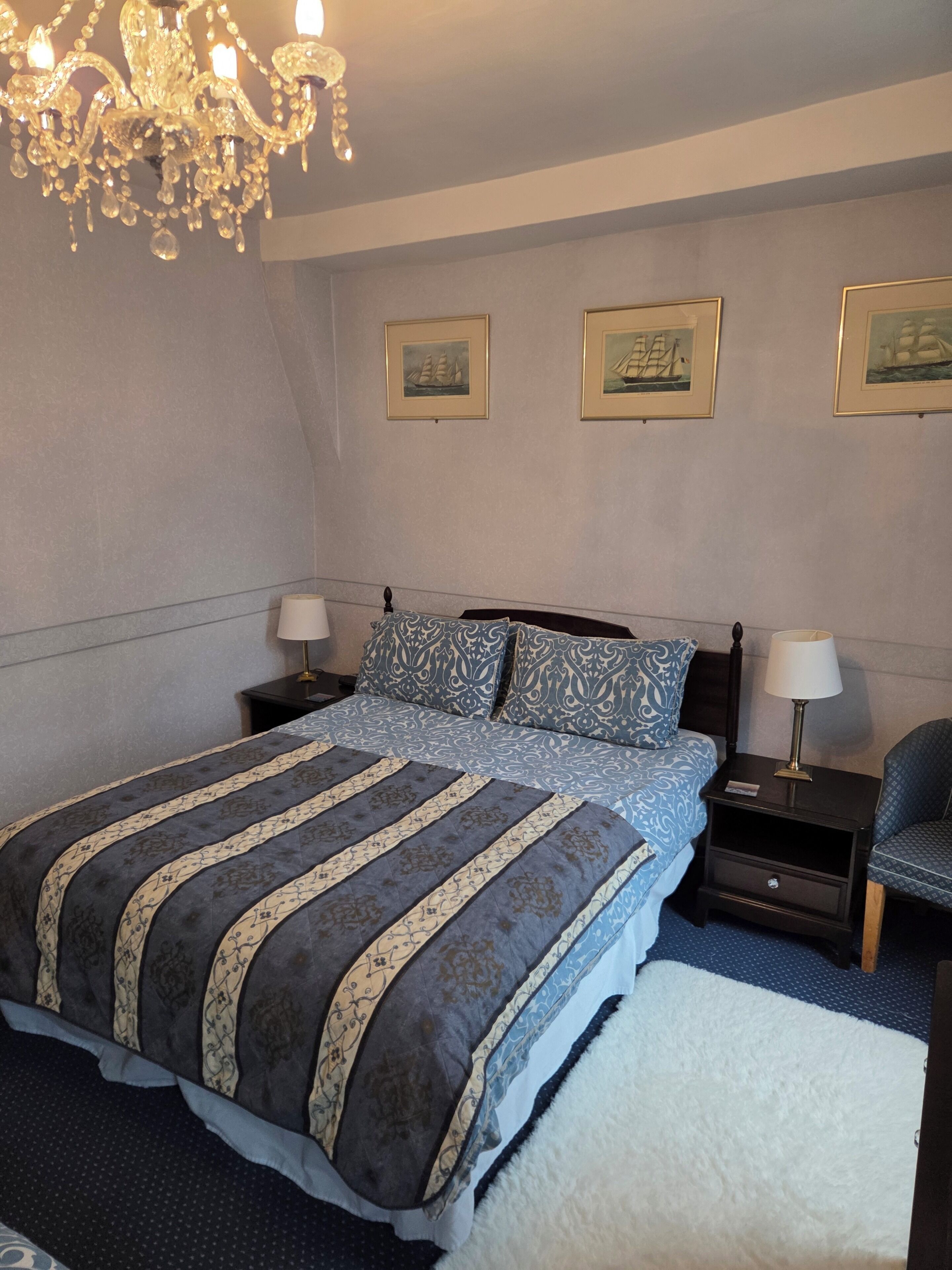 A bedroom with a large bed, two bedside tables, a chair, a chandelier, and three framed pictures on the wall.