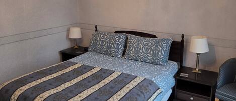 A bedroom with a large bed, two bedside tables, a chair, a chandelier, and three framed pictures on the wall.