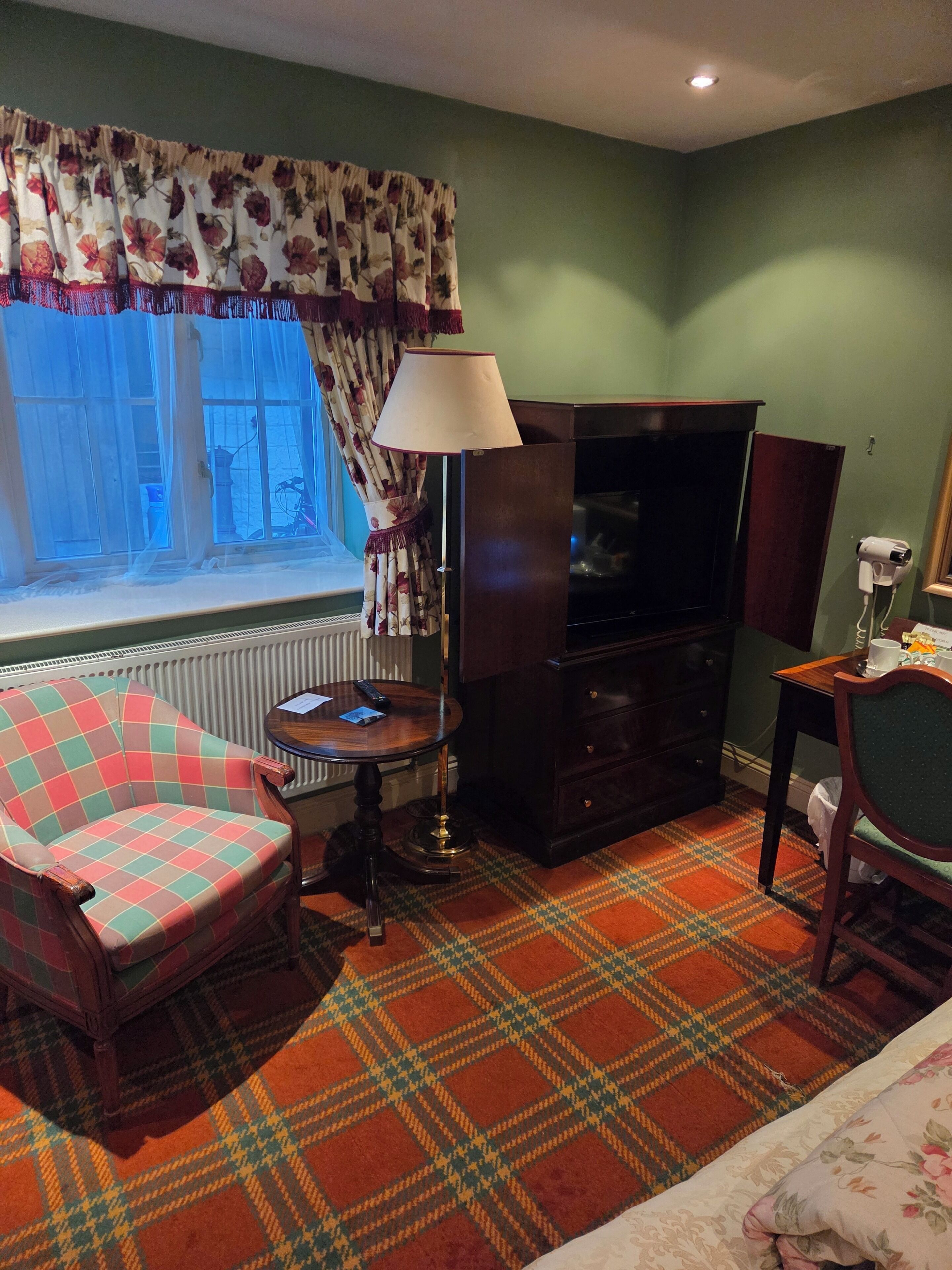 Habitación de hotel con alfombra escocesa, cortina estampada, televisor, lámpara, silla y una pequeña mesa.