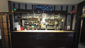 Ein Barbereich mit einem Neonzeichen, das "LANGPORT ARMS!" zeigt, sowie eine Auswahl an Spirituosenflaschen und Gläsern, die in Regalen präsentiert werden.
