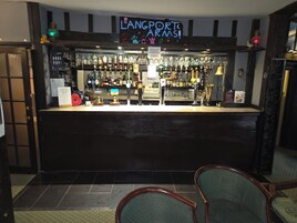 Zona de bar con un letrero de neón que dice "LANGPORT ARMS!" y una variedad de botellas de licor y vasos exhibidos en estanterías.