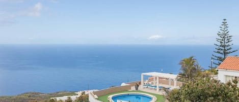 Una piscina con vista al océano, rodeada de vegetación y un edificio con tejado rojo.
