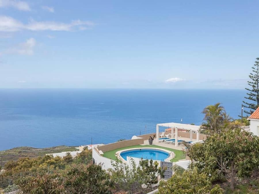 A pool with a view of the ocean, surrounded by greenery and a building with a red roof.