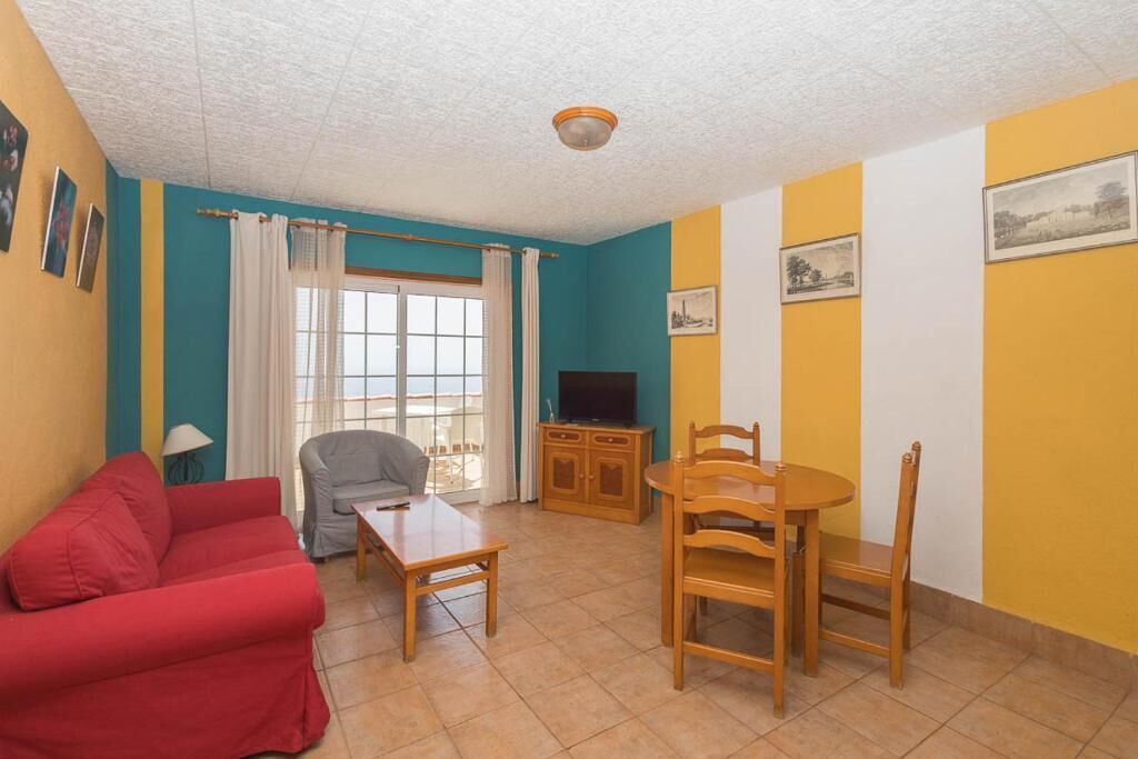Ein Wohnzimmer mit rotem Sofa, grauem Sessel, Esstisch aus Holz und Stühlen. Ein Fernseher steht auf einem Holzschrank, und ein Fenster ist mit Vorhängen drapiert.