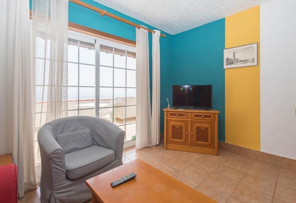 Sala de estar con un sofá gris, un mueble de televisión de madera, un control remoto sobre una mesa y un balcón con cortinas blancas.