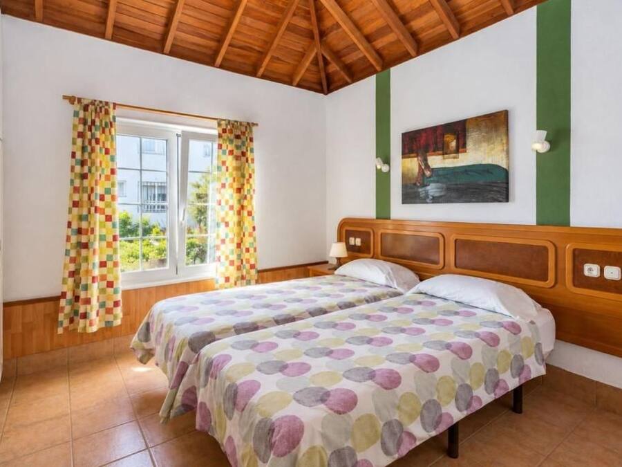 A room with two beds, a wooden ceiling, a window with curtains, and a painting on the wall.