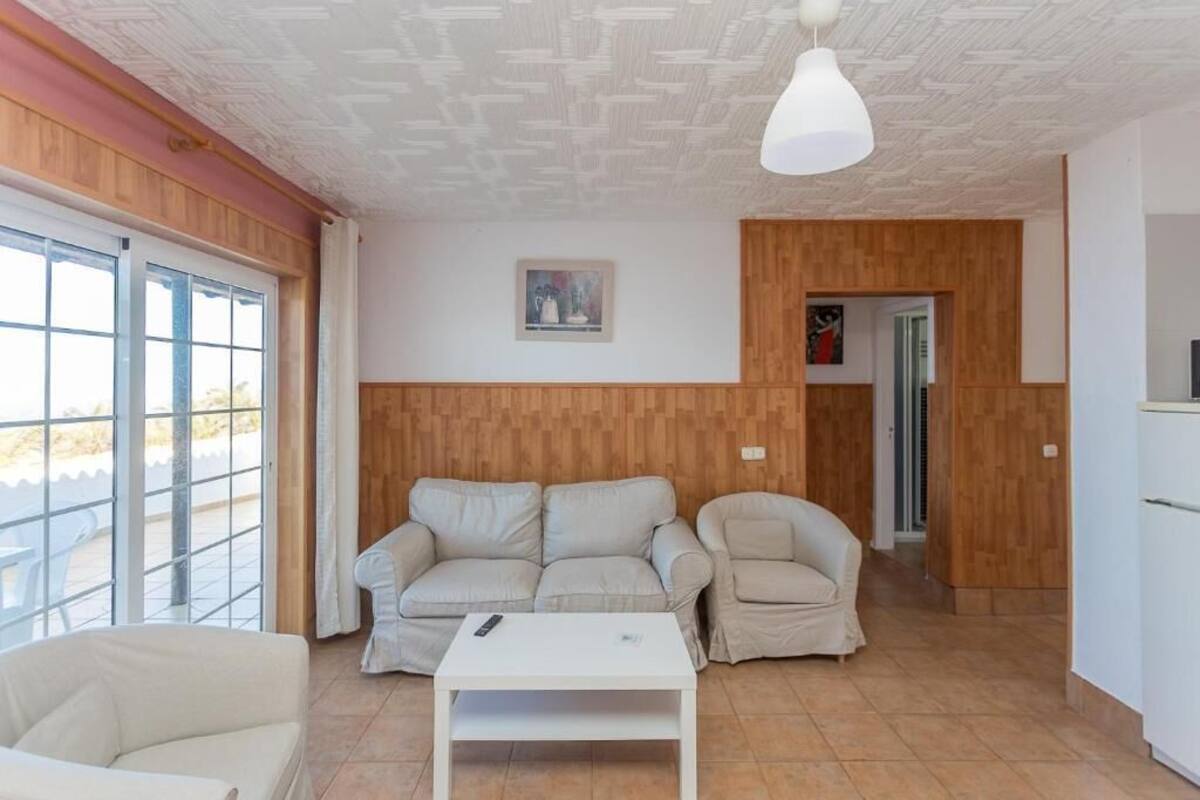 Una sala de estar con un conjunto de sofá, una mesa de centro y un televisor. Hay una puerta que conduce a otra habitación y una ventana con persianas.