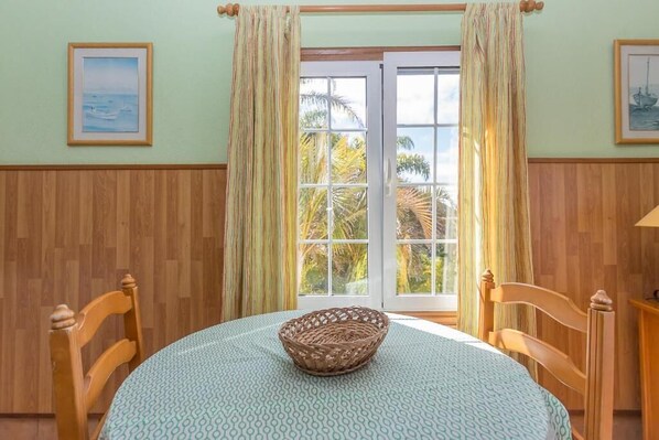 Una habitación con una mesa redonda, dos sillas de madera, una cesta de mimbre, un mantel estampado, una ventana con cortinas y cuadros en las paredes.