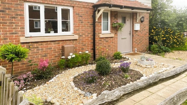 Une maison en briques avec une porte d’entrée, un petit porche et un jardin où poussent différentes plantes et fleurs.