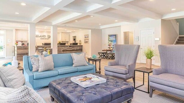 Uma sala de estar espaçosa com um sofá azul, poltronas cinzentas e um banquinho de couro. Uma cozinha é visível ao fundo, com uma placa indicando "ISLAND IS RESORT".