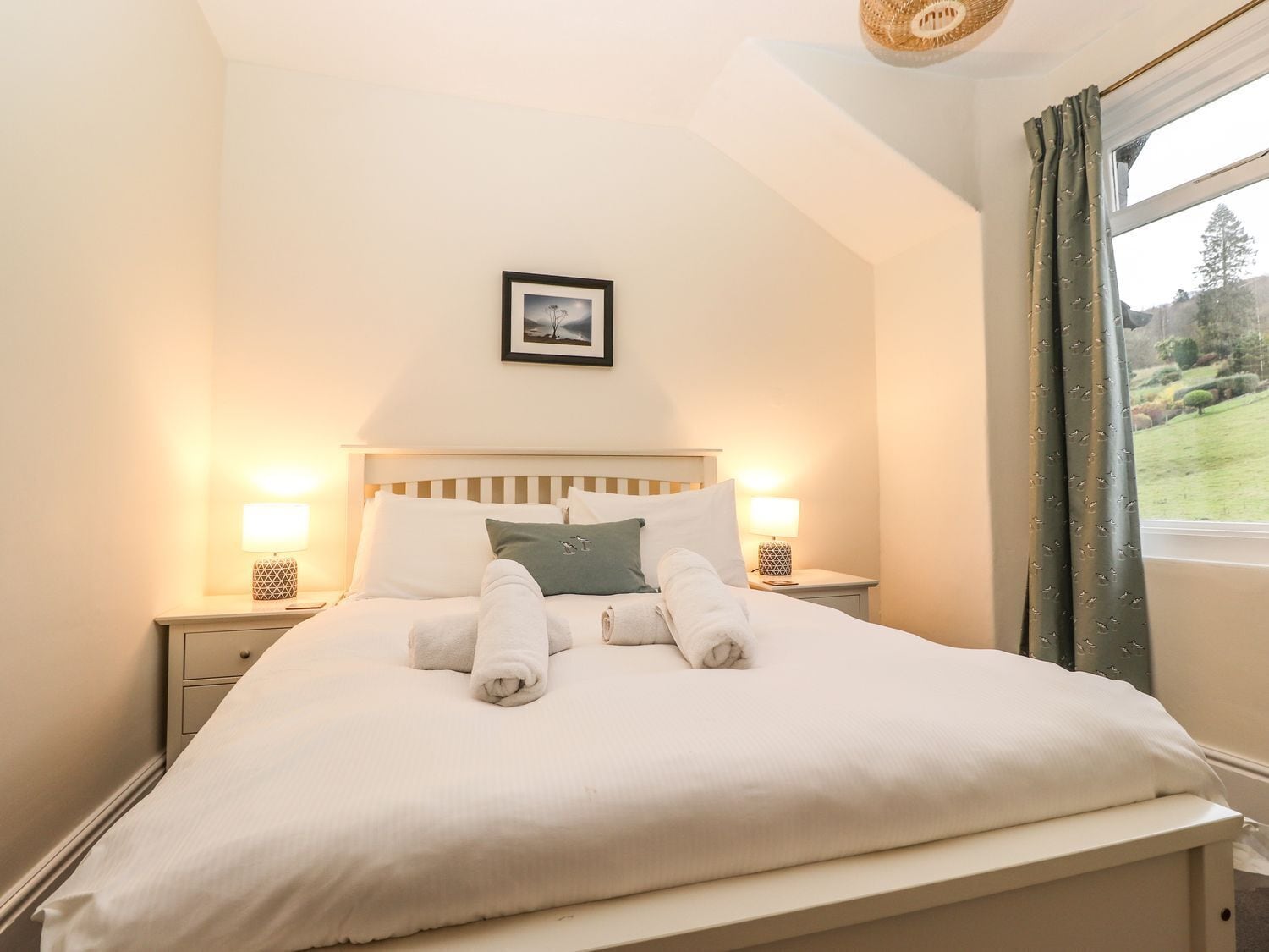 A neatly made bed with white linens and two rolled towels, a framed picture on the wall, and a window with curtains.